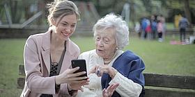 Junge Frau sitzt mit Seniorin auf Bank und zeigt ihr etwas auf dem Smartphone.