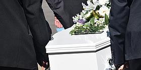 Personen in schwarzen Anzügen tragen einen weißen Sarg auf welchem weiße Blumen sind