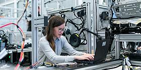 Das Bild zeigt eine junge Frau mit einem Laptop in einer Werkstatt.