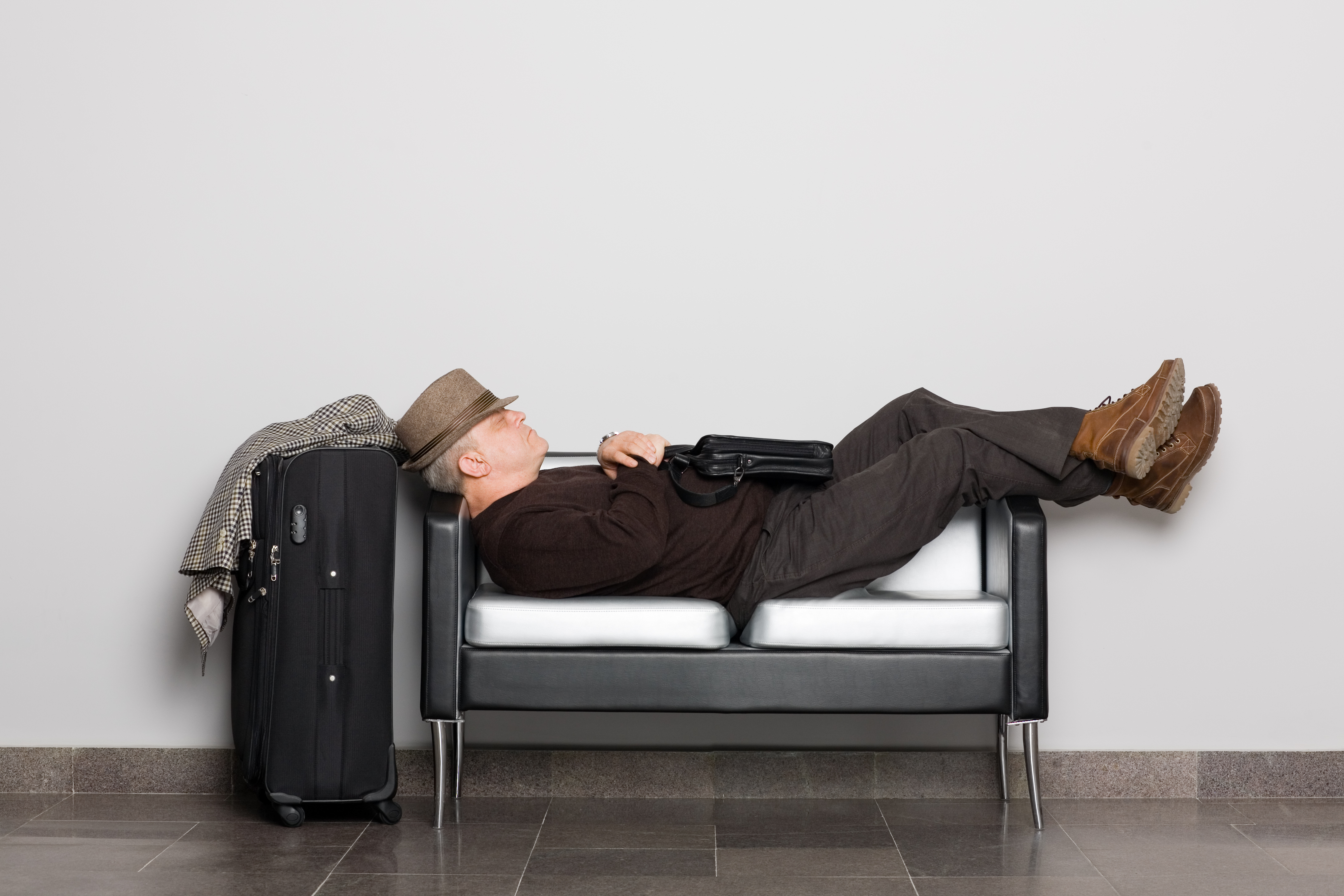 Mann der auf einer kleinen Couch schläft mit Koffer neben der Couch, weil er kein Hotel in der Nähe des Flughafen Wiens gefunden hat.