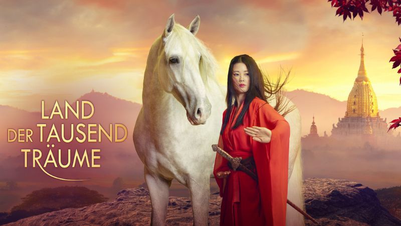 Eine Frau in rotem Gewand und ein weißes Pferd stehen vor malerischer Kulisse.