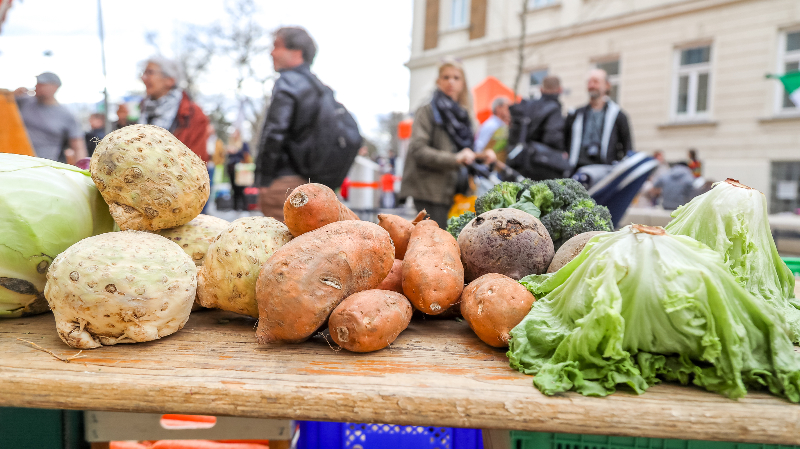 Frisches Gemüse im Vordergrund, dahinter Menschen die einen Markt besuchen