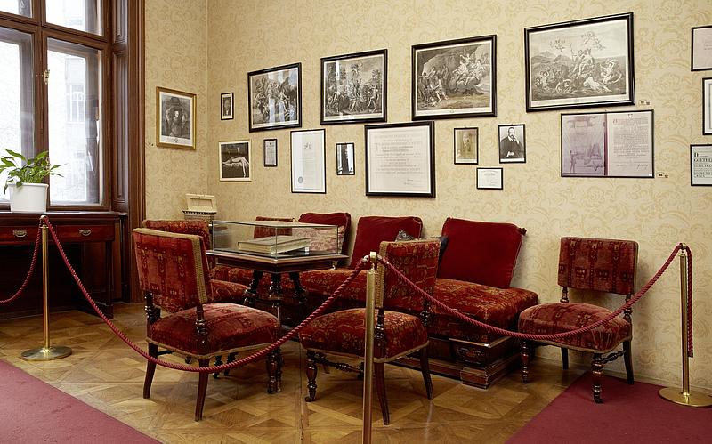 Raum mit vielen samtbezogenen Stühlen, einen kleinen Tisch und vielen Bilderrahmen an der Wand.