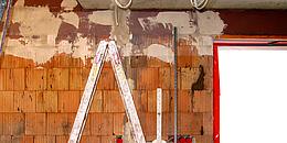 Rohe Ziegelwand in Arbeit, davor steht eine Metallleiter, oben hängen Kabel aus der Decke, seitlich ist ein Fenster ohne Rahmen und Glas