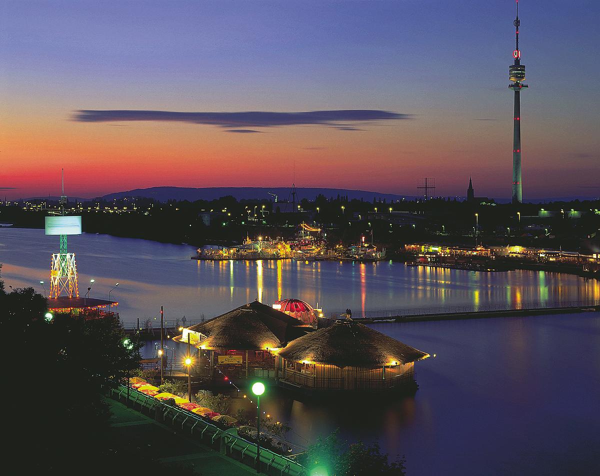 Beleuchteter Donauturm bei Nacht. Im Vordergrund sieht man die "Sunken City" und ihre Lokale.