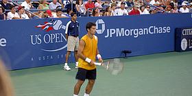 Ein Tennisspieler mit gelbem Shirt, schwarzen Shorts und weißen Schuhe häkt seinen Schläger