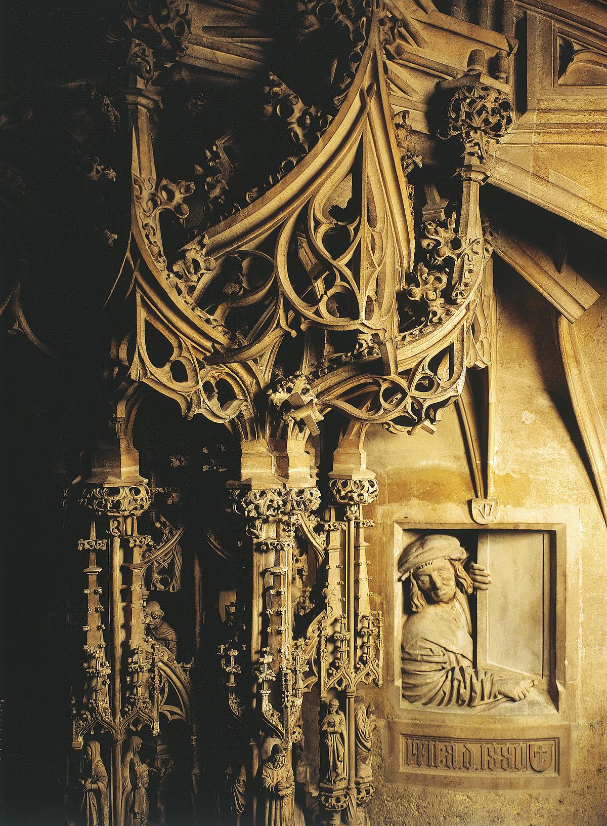 Nahaufnahme von gotischer Kanzel und einer Figur die darunter aus dem "Fenster" sieht.