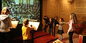 5 junge Kinder dirigieren vor einer Leinwand, auf der ein Konzert abgespielt wird