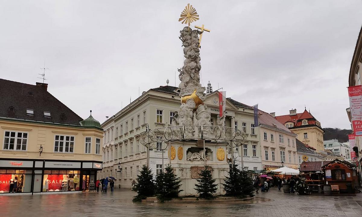 Die Dreifaltigkeitsstatue am Hauptplatz von Baden, umringt von kleinen Tannen, während das Rathaus im Hintergrund zu sehen ist.