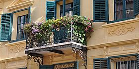 Ein schmiedeeiserner Balkon (an einem dem Baustil nach zu urteilen älter anmutendem Haus) voll mit Blumentrögen und bunten Blüten.