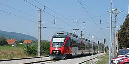 S Bahn-Linie S7 fährt Richtung Floridsdorf, über Flughafen Wien Schwechat. Neben den Gleisen befinden sich Parkplätze.