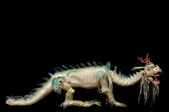 Spielpuppe on der Form eines Drachens mit langem Schwanz, hellem Körper, blauen Rückenspitzen und einem geöffnetem Maul mit spitzen Zähnen.