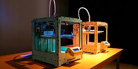 Man sieht zwei würfelförmige 3D-Drucker, wobei im ersten gerade ein Werkstück entsteht.