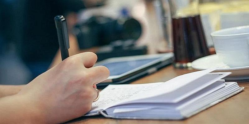 Hand im Fokus mit Stift beim Schreiben in Notizbuch im Hintergrund Kaffeetasse und Laptop