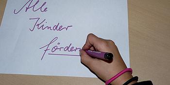Ein Kind schreibt auf ein weißes Blatt Papier die Worte "Alle Kinder fördern"