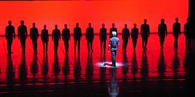 Tanzcompany, auf in rotes Licht getauchter Bühne, mit Choreograf im Vordergrund. 