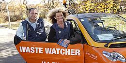 Zwei Personen, die für Waste Watcher arbeiten steigen in ein oranges Auto ein, auf dem "Waste Watcher im Einsatz steht".