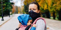 Das Bild zeigt eine Frau mit Gesichtsmaske und ein Baby in einer Babytrage.