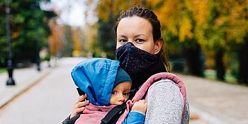 Das Bild zeigt eine Frau mit Gesichtsmaske und ein Baby in einer Babytrage.