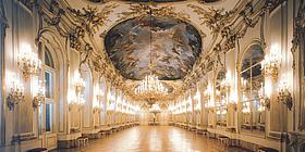 Innenansicht des Schlosses Schönbrunn: die prunkvolle Große Galerie wird von einem großen Kronleuchter in der Mitte und von vielen kleinen am Gang hell erleuchtet. 