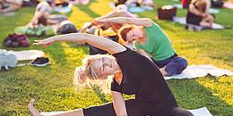 Bewegt im Park Symbolbild: Im Vordergrund Frau auf Yogamatte sitzend in Stretch-Position auf einer Wiese, im Hintergrund weitere Menschen in derselben Position