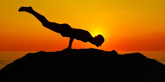 Person, die die Yoga Figur Pfau auf einem Felsen vor der untergehenden Sonne macht 