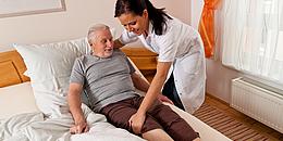 Pflegerin hilft älterem Mann, der in einem Bett liegt.