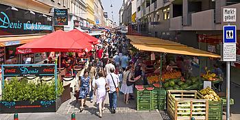Bild vom Eingang des Brunnenmarktes: Lokale und Verkaufsstände auf beiden Seiten, letztere bieten frisches Obst und Gemüse an.