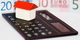 Ein kleines Hausmodell mit rotem Dach steht auf einem Taschenrechner, mit Euroscheinen im Hintergrund.
