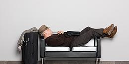 Mann der auf einer kleinen Couch schläft mit Koffer neben der Couch, weil er kein Hotel in der Nähe des Flughafen Wiens gefunden hat.