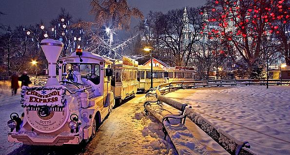 Ein kleiner Expresszug fährt durch einen schneebedeckten Park, während an Bäumen und Gebäuden bunte Lichter leuchten