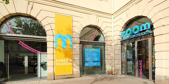 Außenansicht eines Gebäudes. Im Zentrum sind zwei Glastüren mit der Schrift ZOOM Kindermuseum.