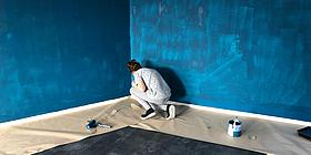 Frau hockt am Boden vor blauer gefleckten Wand und streicht sie