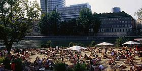 Viele Menschen sitzen auf Liegestühlen unter >Sonnenschirmen am Strand vorm Donaukanal