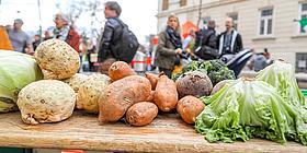 Frisches Gemüse im Vordergrund, dahinter Menschen die einen Markt besuchen