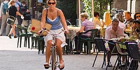 Frau am Fahrrad in der Stadt dahinter Leute im Gastgarten