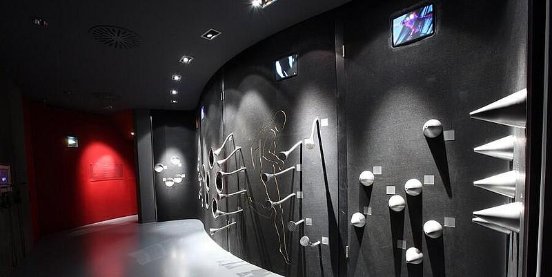 Raum in grau mit rotbeleuchtetem Ausgang und Lautsprecherrohren an der Wand als Klanggalerie