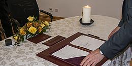 Eheschließund auf dem Standesamt: Auf einem Tisch mit weißer Tischdenke, Blumenkranz und einer brennenden Kerze sieht man die Trauurkunde