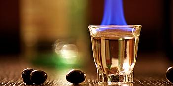 Hochprozentiger Alkohol in einem Glas, von dem eine blaue Flamme aufsteigt umd um welches herum Oliven liegen 