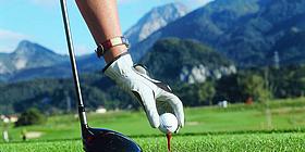Eine Hand hält den Golfball, daneben ein Golfschläger. Im Hintergrund sieht man die Alpen.