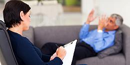 Therapeutin im Vordergrund mit Notizblock und Patient im Hintergrund auf einer Couch