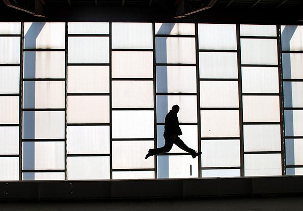 Bild zeigt einen Mann in der Luft schwebend vor dem Hintergrund aus rechteckigen Fenstern mit schwarzen Rahmen.