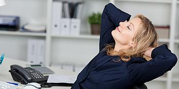 Frau die entspannt auf einem Bürosessel sitzt und die Arme hinter dem Kopf verschränkt hält