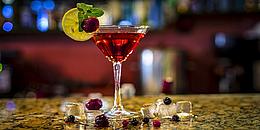 Martiniglas mit rotem Cocktail auf Bar, darutner liegen Eiswürfel und Himbeeren.