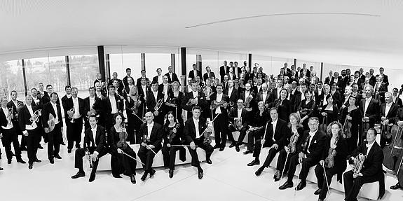 Orchester der Symphoniker in Wien abgebildet schwarz - weiß, im Vordergrund sieht man sitzende Musiker mit ihren Instrument die anderen stehen im Hintergrund