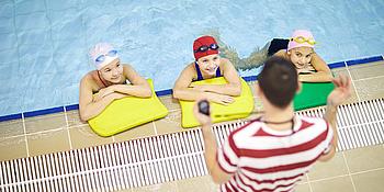 Schwimmkurs für Kinder, drei Kinder im Wasser und der Schwimmlehrer steht am Beckenrand