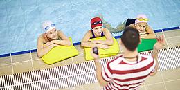 Schwimmkurs für Kinder, drei Kinder im Wasser und der Schwimmlehrer steht am Beckenrand