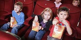 Drei Kinder sitzen in roten Kinositzen und essen dabei Popcorn.