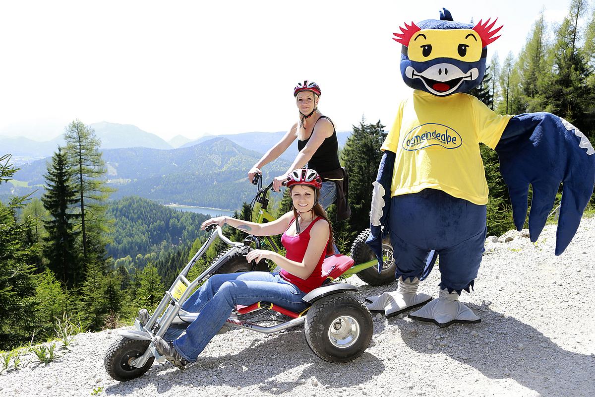Maskottchen der Gemeindalpe mit zwei Frauen, die auf Mountaincart und Monsterroller sitzen