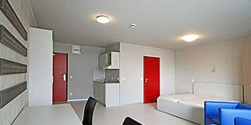 Pärchen-Appartment im Studentenwohnheim base 11, rote Einganstür und rote Türe zum Badezimmer, Raum ansonsten in hellem Weiß gehalten. weißes modernes Doppelbett, kornblaue Sitzmöbel. Doppelschreibtisch mit schwarzen Bürosesseln.